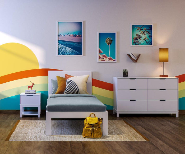 A colorful bedroom features a white bed with blue bedding and a yellow backpack. Rainbow stripes and a sun are painted on the wall above three framed photos that hang over the bed and dresser. The modern Lanson V Shelf adds geometric flair beside solid poplar shelves, while a lamp and decor sit atop the dresser and nightstand.