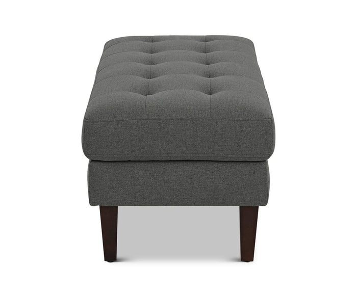 The Laura Ottoman is a rectangular piece upholstered in dark gray fabric, featuring a tufted top and dark wooden legs; this modern ottoman is displayed from the front against a white background.