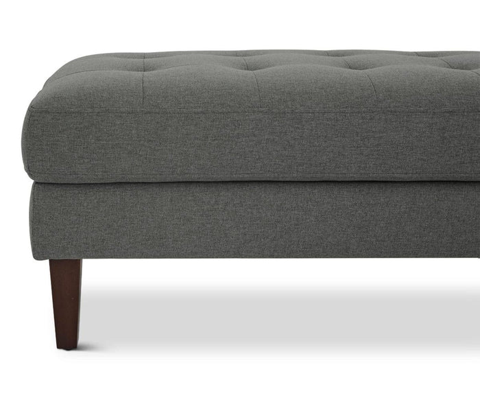 A close-up of the Laura Ottoman, featuring gray upholstery, tufted buttons, and dark wooden legs, displayed against a white background.