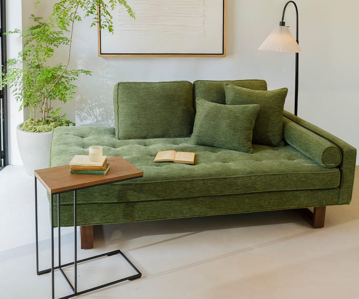 A green daybed with two cushions and a bolster sits near a potted plant. Next to it, the Leende C-Table, crafted from European oak, holds books and a candle. A floor lamp stands beside framed artwork hanging on the wall behind the daybed, creating a calm and cozy interior.
