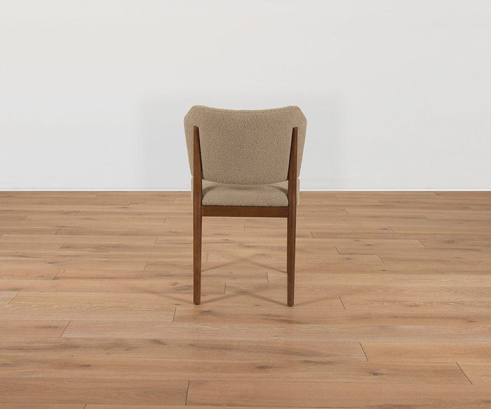 A Leevi Dining Chair, featuring a beige cushion, solid rubberwood frame, and wooden legs, is turned backward on a light wooden floor against a plain white wall.