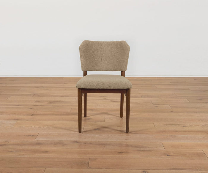 A Leevi Dining Chair featuring a mid-century design with tan performance fabric upholstery on the seat and backrest, and solid rubberwood legs, is positioned on a light brown hardwood floor in front of a plain white wall.