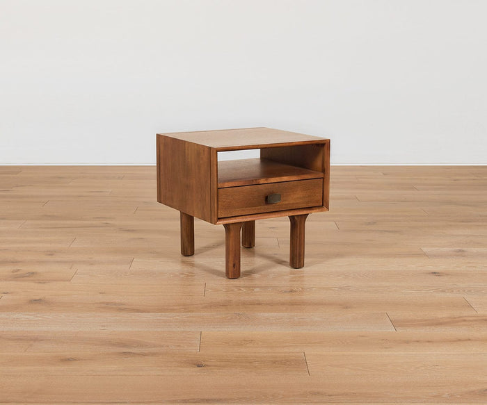 The Leevi End Table, expertly crafted from solid acacia wood, features four legs, an open top shelf, and a single drawer with a metal handle. This mid-century piece from the Leevi Collection is shown on a light wood floor against a white wall.