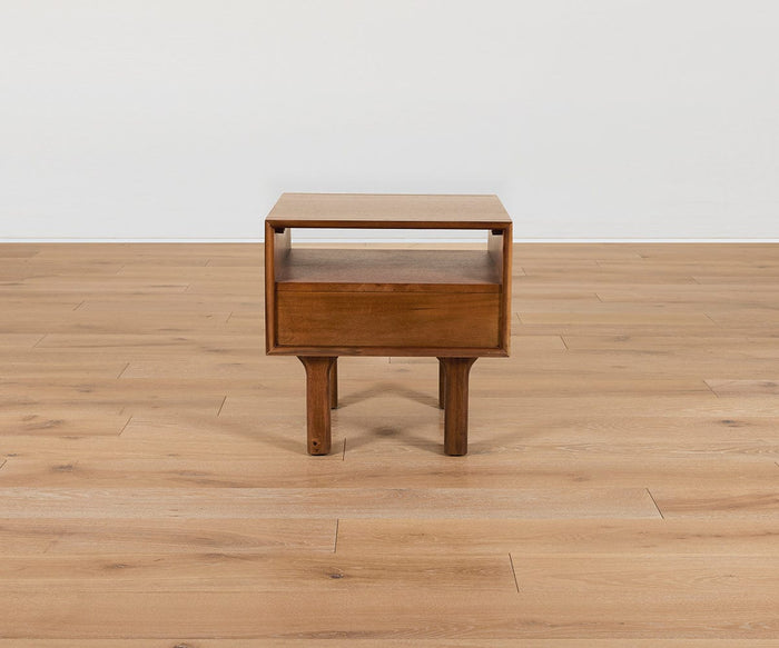 The Leevi End Table—a small, mid-century piece crafted from solid acacia wood—features an open shelf and a drawer, standing on four legs atop a light wood floor against a plain white wall.