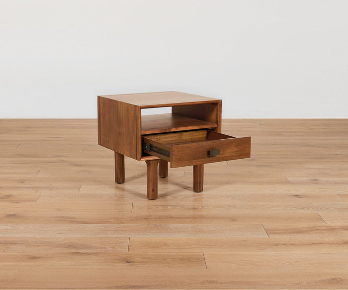 The Leevi End Table from the Leevi Collection is expertly crafted from solid acacia wood and showcases four sturdy legs, an open lower shelf, and a single drawer that is partially pulled out. This elegant mid-century piece sits on a light wood floor against a white wall.
