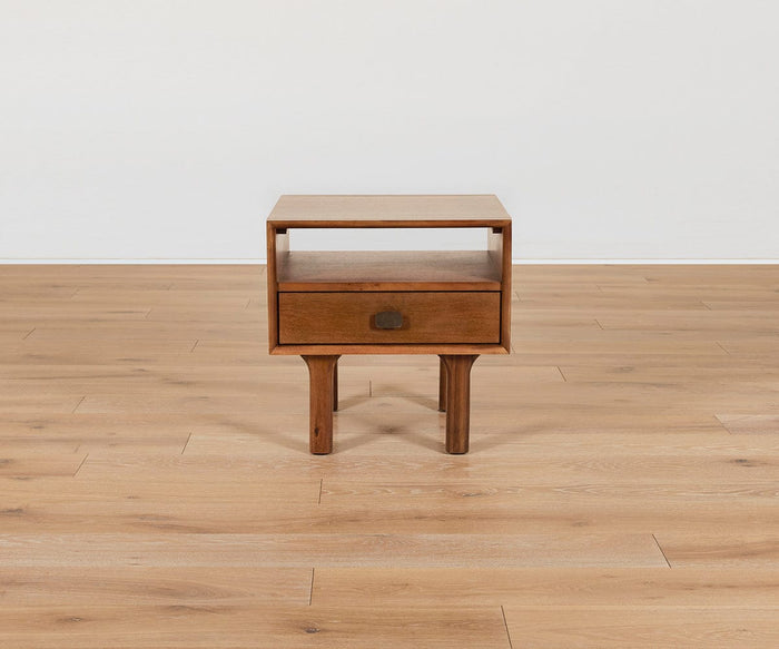 The Leevi End Table, crafted from solid acacia wood with one drawer and an open shelf, stands on four legs atop a light wood floor against a plain white wall.