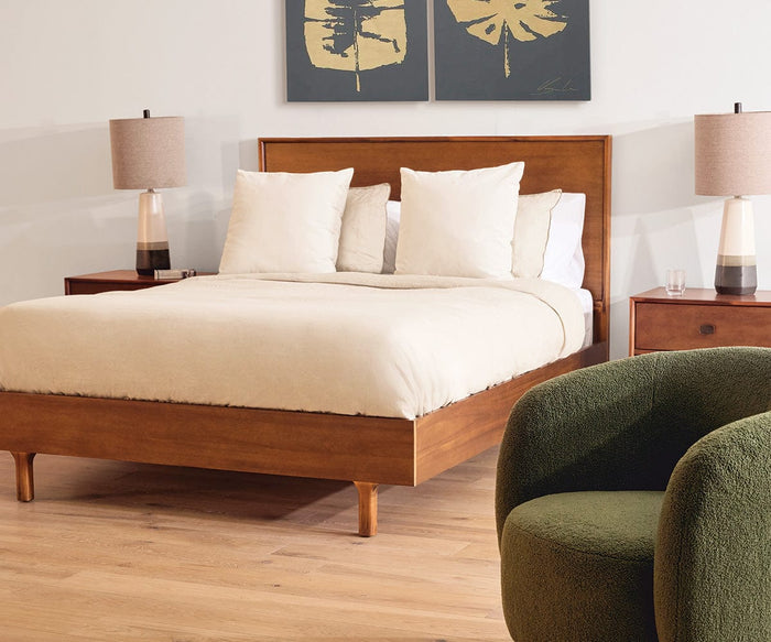 A Leevi Bed, crafted from solid acacia wood and dressed in crisp white bedding with pillows, is flanked by two nightstands topped with matching lamps. A green chair sits in the foreground, while a pair of botanical prints displayed above the bed enhance its timeless mid-century design.