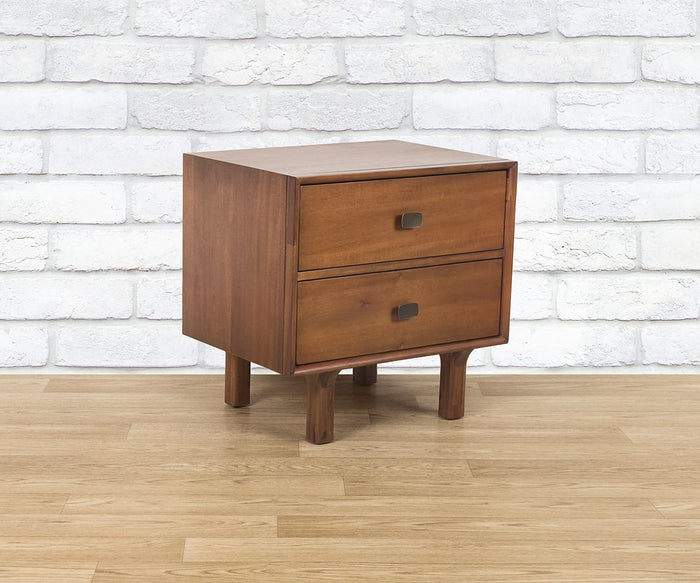 The Leevi Nightstand, expertly crafted from solid acacia wood, showcases two spacious drawers with sleek square handles and is supported by four sturdy legs. Placed before a white brick wall and atop light wood flooring, this mid-century piece brings a touch of timeless elegance to any bedroom setting.