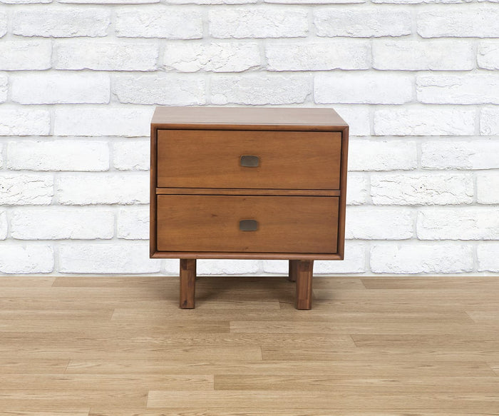 The Leevi Nightstand, crafted from solid acacia wood, features two drawers adorned with square metal handles and stands elegantly on a light wood floor against a white brick wall. This stylish mid-century piece seamlessly blends timeless design with modern functionality.