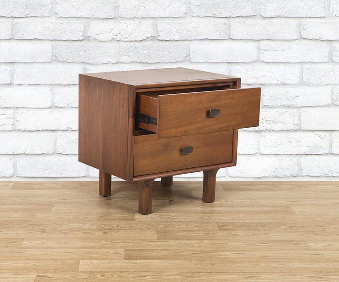 The Leevi Nightstand, crafted from solid acacia wood, features two drawers—with one shown partially open—and stands on four sturdy legs. It is placed on a wooden floor with a white brick wall as the backdrop.