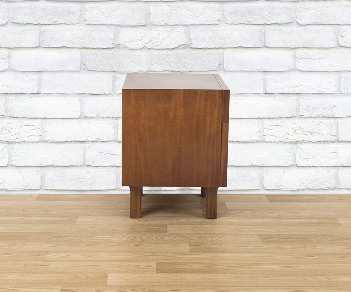 A Leevi Nightstand stands on a light wood floor in front of a white brick wall, showcasing its solid acacia wood construction from a side perspective.