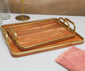 The Leon Trays - Set of 2, crafted in a rich cherry veneer and adorned with elegant gold-toned handles, are stacked on a white surface beside a pink napkin and assorted glass and brass objects.