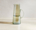 A tall, clear Lunna Glass Pitcher with a handle is half-filled with water and sits on a light-colored surface against a neutral background. This dishwasher safe pitcher in glass features a subtle ribbed luster finish, adding an elegant touch to any setting.