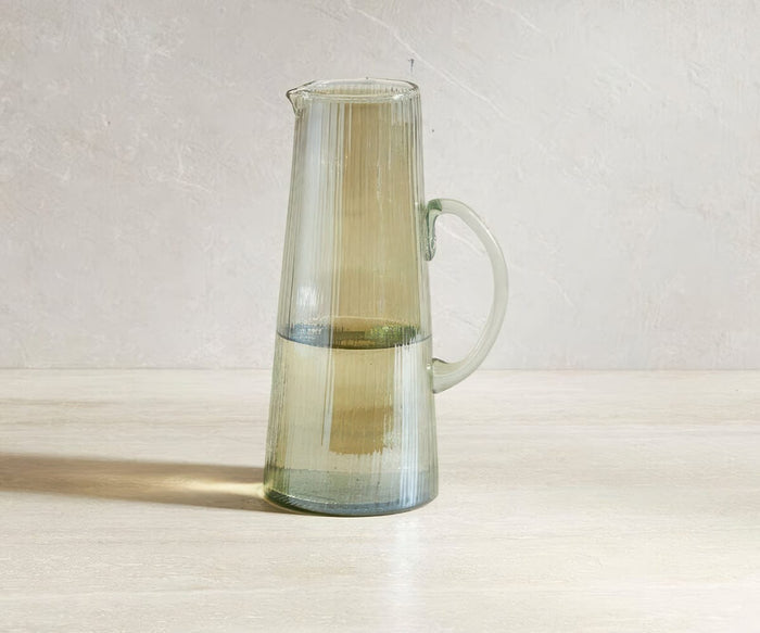 A tall, clear Lunna Glass Pitcher with a handle is half-filled with water and sits on a light-colored surface against a neutral background. This dishwasher safe pitcher in glass features a subtle ribbed luster finish, adding an elegant touch to any setting.