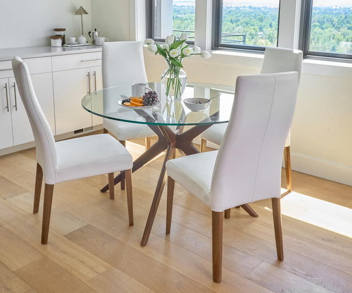 A glass dining table paired with four Lussa Leather Dining Chairs, each featuring elegant leather upholstery and solid wood legs, accompanied by a vase of white flowers, a bowl, and a plate of fruit in a bright room with large windows and wooden floors.