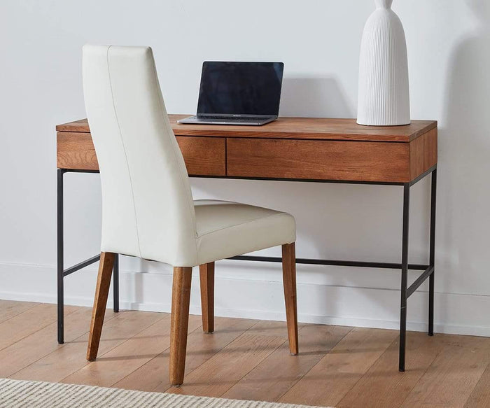 A wooden desk with black metal legs, a closed laptop, a tall white vase, and a Lussa Leather Dining Chair are set up in a minimalist room with a light wood floor.