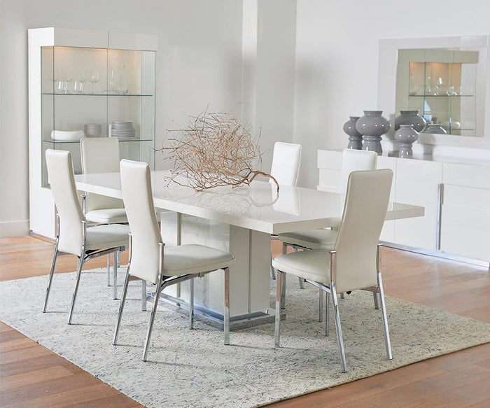 A modern dining room with a white table, six Lykke High Back Dining Chairs that offer padded comfort, a glass cabinet with dishes, a white sideboard with gray vases, a decorative branch centerpiece, and a light area rug on wooden floors.