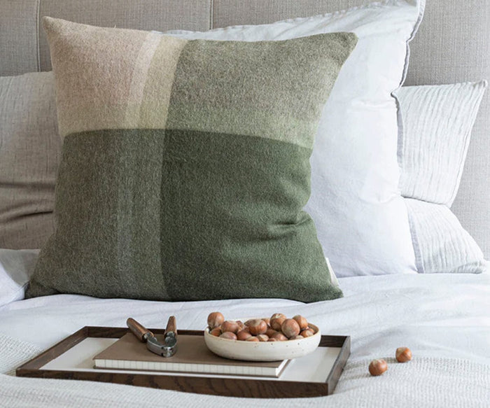 A large, square Manhattan Cushion Cover in green and beige color blocks rests on a neatly made bed, its fabric crafted from recycled alpaca wool for a soft, sustainable touch. In front, a wooden tray holds a bowl of hazelnuts, a nutcracker, and a notebook.