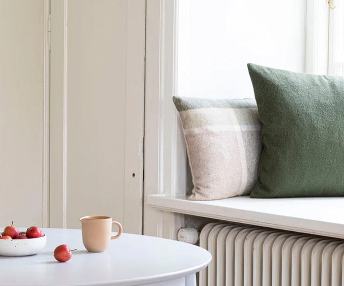 A white table with a beige mug and a bowl of small red apples sits near a window seat adorned with Manhattan Cushion Covers in green and beige, crafted from recycled alpaca wool. Light streams in from the window above a white radiator, highlighting the Fair Trade textiles that bring warmth to the cozy nook.