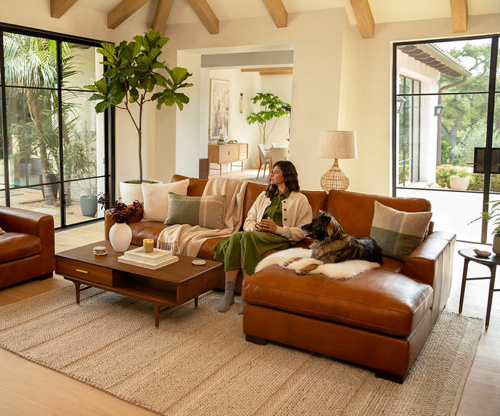 A woman in a green dress sits on a brown leather sofa with a dog beside her in a bright, modern living room featuring large windows, plants, and neutral decor, accented by the Manhattan Cushion Cover.