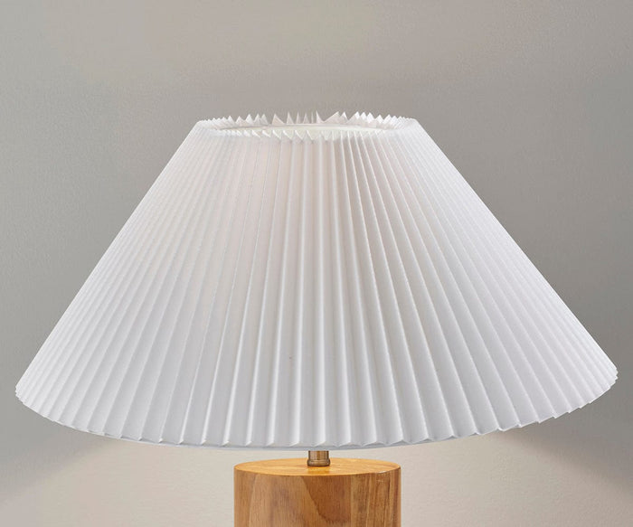 The Marta Table Lamp features a white pleated lampshade atop a natural wood base, creating a modern yet timeless accent. It stands out beautifully against the plain light gray wall in the background.