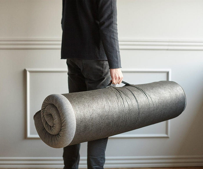 A person dressed in dark clothing holds a rolled-up Mattress Topper with a handle in a light-colored room featuring decorative wall molding.