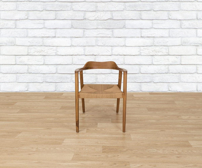 A Melina Chair is centered and facing the camera, featuring a woven seat and crafted from solid teak. It is positioned on a light wood floor in front of a white brick wall.