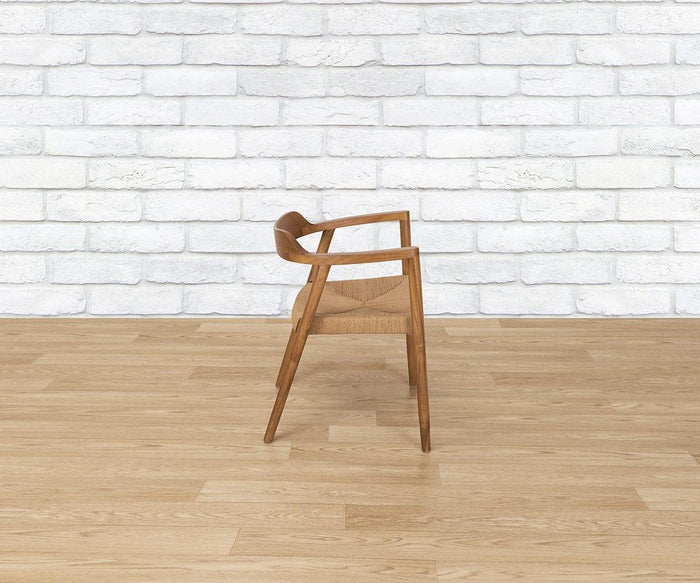 The Melina Chair, featuring angled wooden legs and a woven seat, is shown from the side, placed on a light wood floor with a white brick wall in the background.