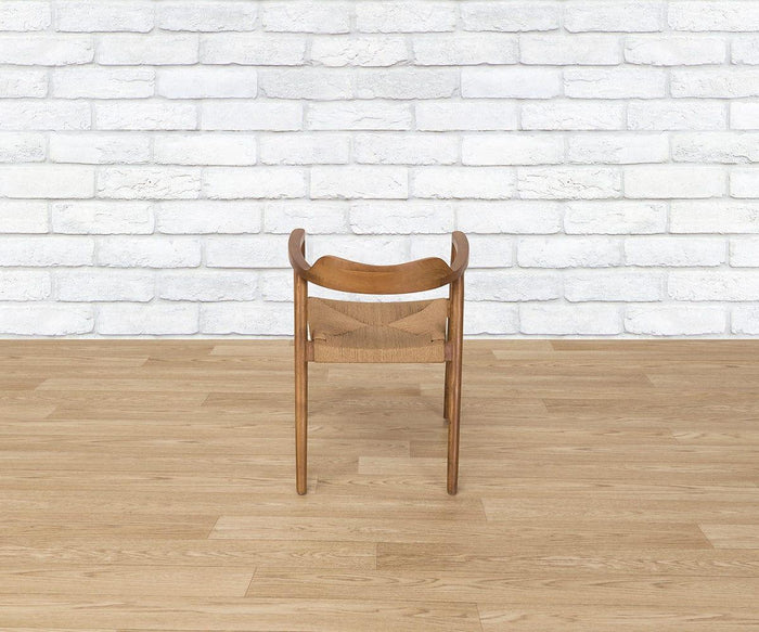 The Melina Chair, featuring a curved seat and woven backrest, is positioned backwards on a light wood floor against a white brick wall.