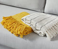 Two folded throws with fringe lie on a light gray sofa. One decorative throw is mustard yellow, while the other is the Minimalist Stripe Throw in white, featuring thin black stripes. Both have a textured, woven appearance.
