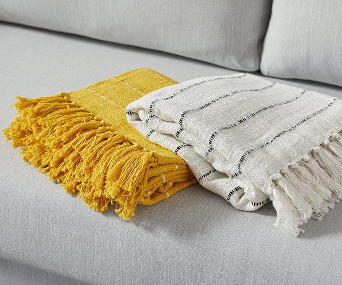Two folded throws with fringe lie on a light gray sofa. One decorative throw is mustard yellow, while the other is the Minimalist Stripe Throw in white, featuring thin black stripes. Both have a textured, woven appearance.