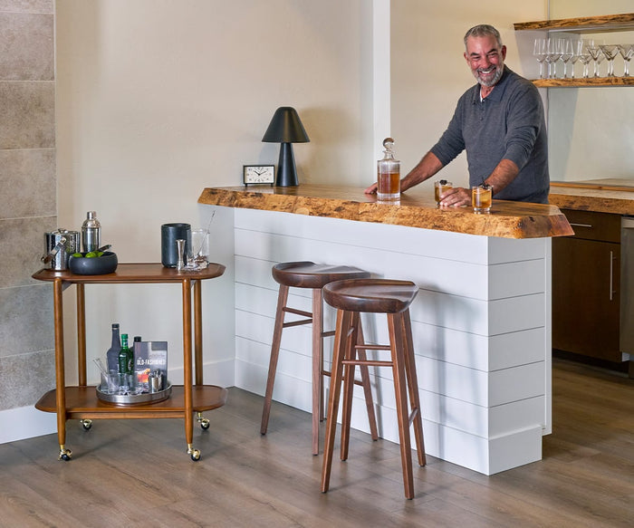 A smiling man stands behind a modern home bar with two wooden stools, the elegant decor enhanced by the Mirco Black Marble Vessel prominently displayed on the sleek bar. A bar cart with liquor and glassware is nearby. The room has wood floors, beige walls, and a small lamp on the bar.