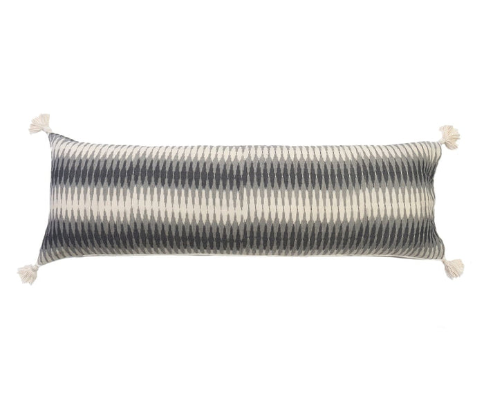 The Miti Bolster Pillow features a rectangular shape with a grey and multi-colored geometric zigzag pattern, crafted from cotton ikat fabric and finished with tassels on each corner for added dimension.