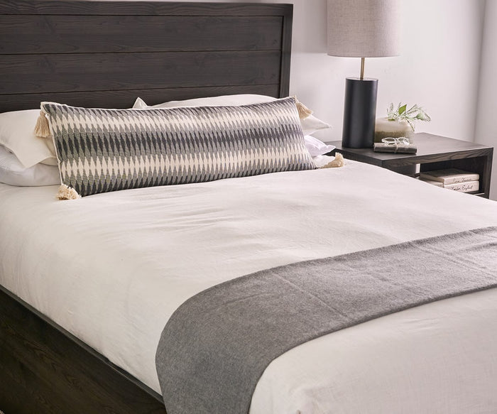 A neatly made bed with white sheets, a gray throw blanket at the foot, the Miti Bolster Pillow in cotton ikat fabric adding a multidimensional look, a dark wooden headboard, and a bedside table with a lamp, book, and small plant.