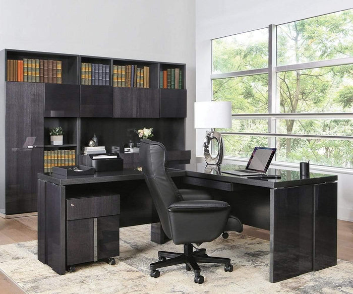 Modern office with a black desk featuring excellent desk organization and the sleek Mondiana Central Keyboard, complemented by an office chair, laptop, lamp, and bookshelves. Large windows let in natural light, offering views of trees outside. A rug rests on the wooden floor. The space is tidy and professional—a perfect blend of style and function made in Italy.