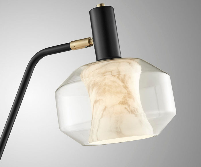The Naeva Table Lamp features a modern double-glass design with a black and gold stem, a clear geometric glass shade, and an inner white marbled diffuser, providing soft illumination against a plain gray background.
