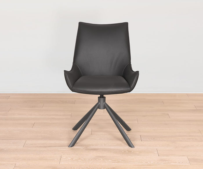 The Nomi Dining Chair is a contemporary piece showcasing a black swivel design, curved backrest, and integrated armrests. Finished with a sleek faux leather seat and supported by a sturdy four-legged metal base, this chair stands gracefully on a light wooden floor in front of a plain white wall.