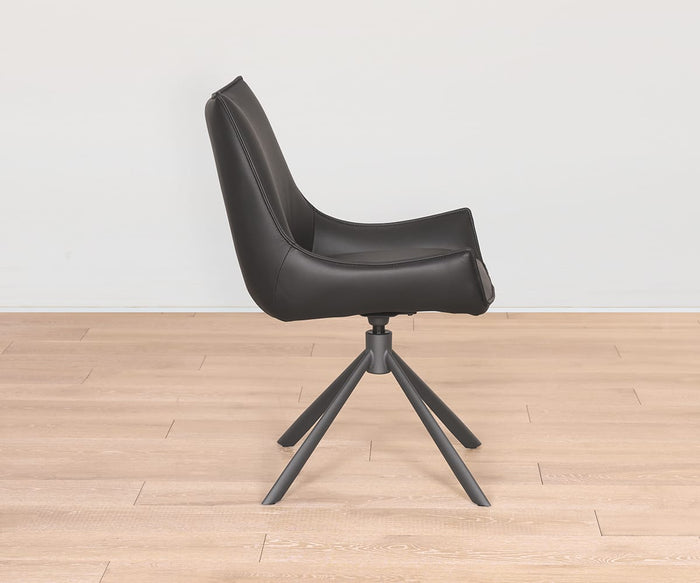 A contemporary Nomi Dining Chair with a black faux leather seat, swivel design, and angled metal legs is shown in side view, positioned on a light wood floor against a plain white wall.