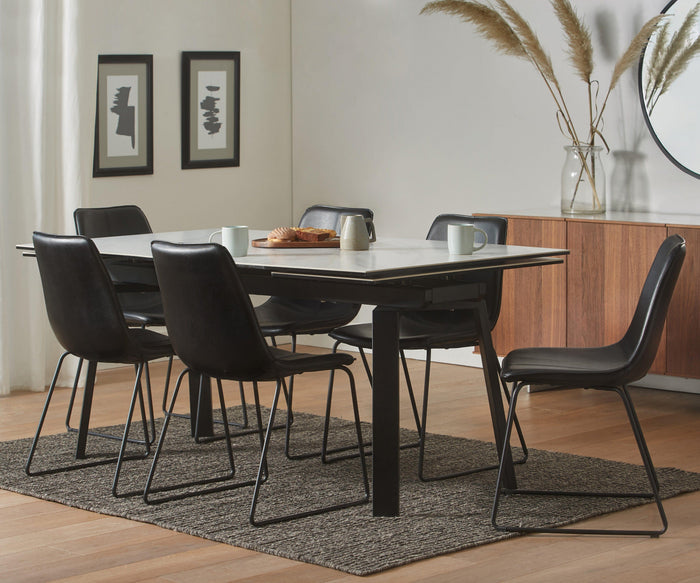 Modern dining room with a rectangular table, six Oken Dining Chairs featuring sleek black frames, a brown rug, wooden sideboard, large round mirror, two framed abstract art pieces, and pampas grass in a vase. Four mugs sit on the table.