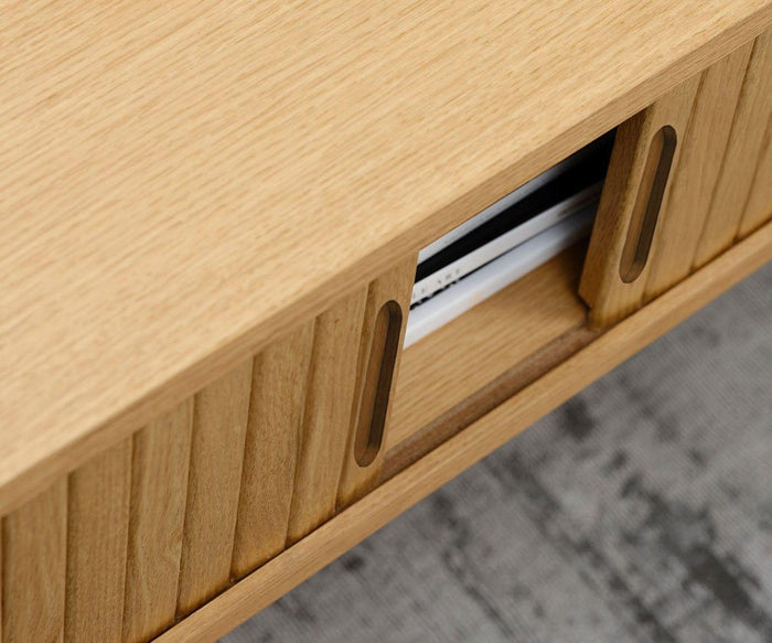 Close-up of the Orella Storage Coffee Table, showcasing its elegant tambour paneling on two sliding wooden doors. One door is partially open, revealing a few magazines or books neatly organized inside—ideal for a modern living room. The coffee table sits gracefully on a grey textured floor.