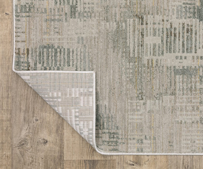 A corner of the Oriental Weaver Mallory / MAL03 Rug, a patterned gray power-loomed area rug, is flipped over to reveal its underside. This earthy rug rests on a naturally finished wooden floor.