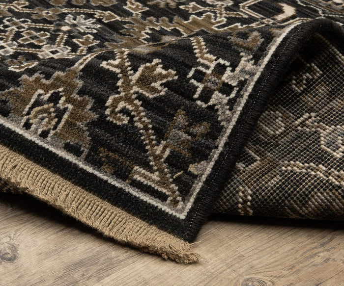 A close-up of a corner of the Oriental Weavers Acacia / ACA05 Rug, featuring black, brown, and beige geometric patterns in a boho style, folded over to show the underside, set on a wooden floor.