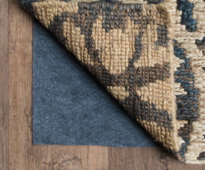 An Oriental Weavers All-N-One / 0004E Rug Pad is partially visible beneath a woven rug that’s lifted in the corner, offering essential floor protection on a wooden floor.
