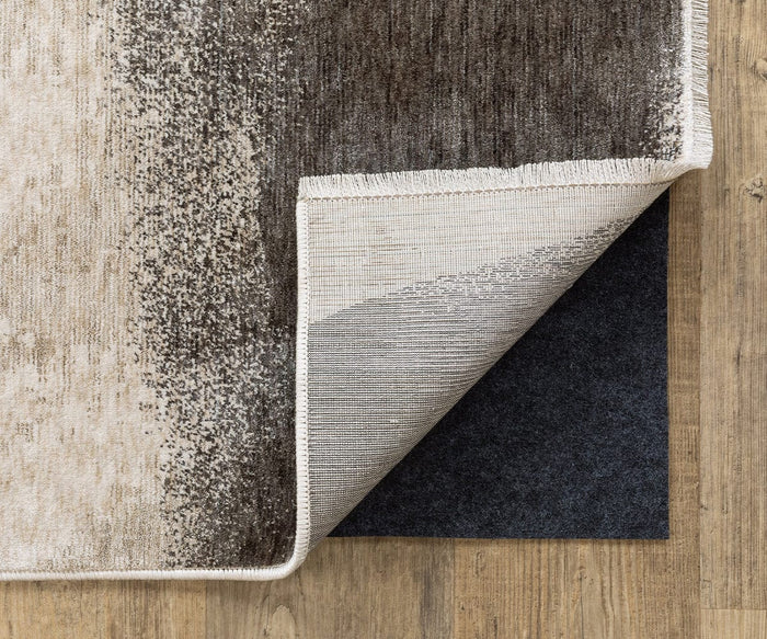 A corner of a beige and brown area rug is lifted to reveal the Oriental Weavers All-N-One / 0004E Rug Pad underneath, providing floor protection on a wooden floor.