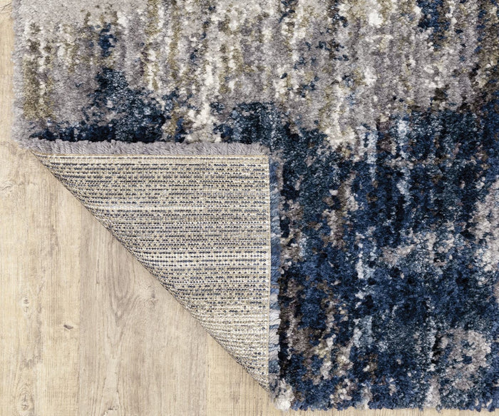 The Oriental Weavers Aspen / 2061L Rug, shown in shades of blue, gray, and white on a wooden floor, features an abstract design crafted from space-dyed microfiber polyester for a richly textured appearance. One corner is folded over to display the woven underside in coordinating neutral tones with a different pattern.