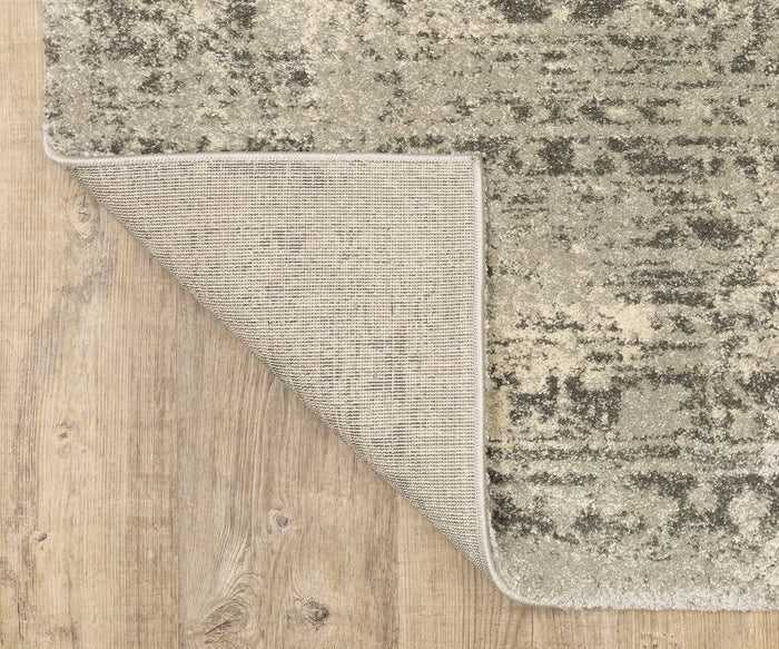 A corner of the Oriental Weavers Astor / 1806Q Rug, showcasing its textured beige and gray design, is flipped over to reveal the woven underside. This plush pile rug, adorned with vintage-inspired motifs, rests on a light wood floor with visible grain patterns.