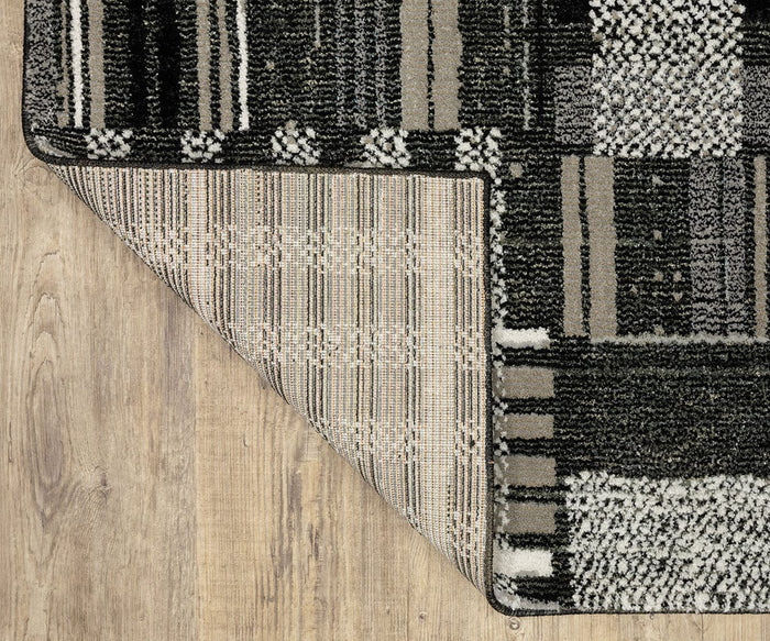 The Oriental Weavers Atlas / 752C Rug, featuring a black, gray, and white pattern, is displayed on a wooden floor with one corner flipped over to reveal its durable machine-woven underside—making it ideal for high-traffic areas.