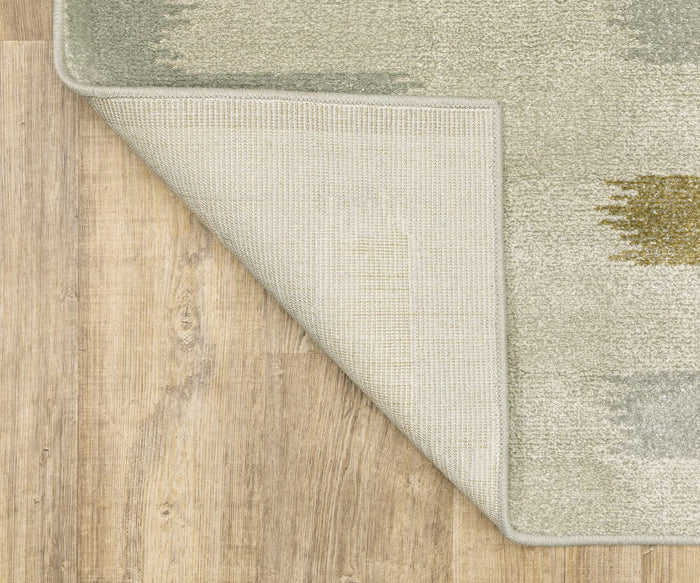 A corner of the light-colored Oriental Weavers Branson / BR09A Rug is flipped over on a wood floor, revealing its underside and textured backing. This power-loomed rug showcases modern motifs that enhance the natural, light brown wood grain pattern below.
