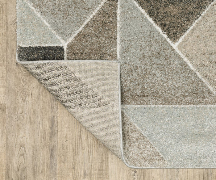 The Oriental Weavers Cambria / 4L Rug, featuring a geometric pattern in neutral tones, is partially folded to reveal its non-slip backing. Made from soft polypropylene yarn, this high-pile rug rests on a light wood floor.