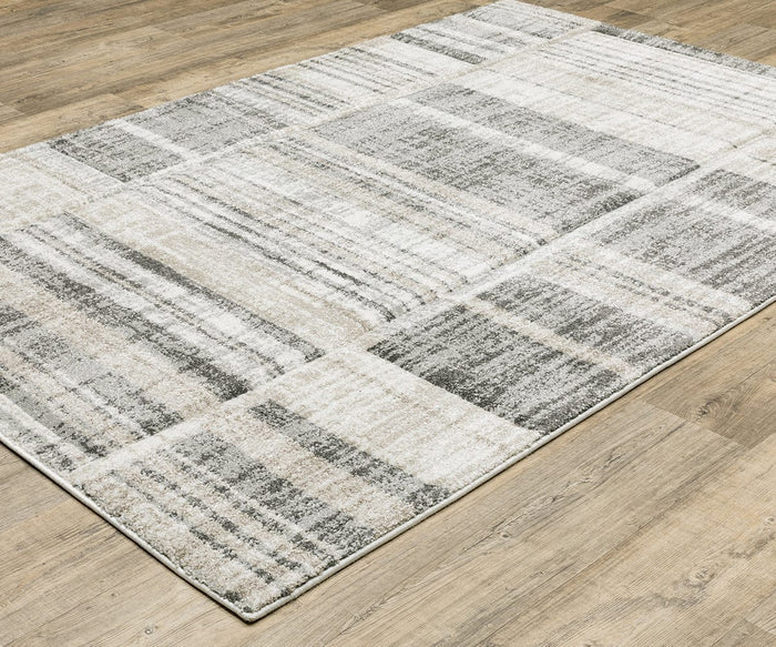 The Oriental Weavers Cambria / 55X Rug, featuring a modern abstract pattern in shades of gray, beige, and white, sits on a light wood floor. Woven from soft polypropylene yarn, this rectangular rug showcases a patchwork design with textured linear details.